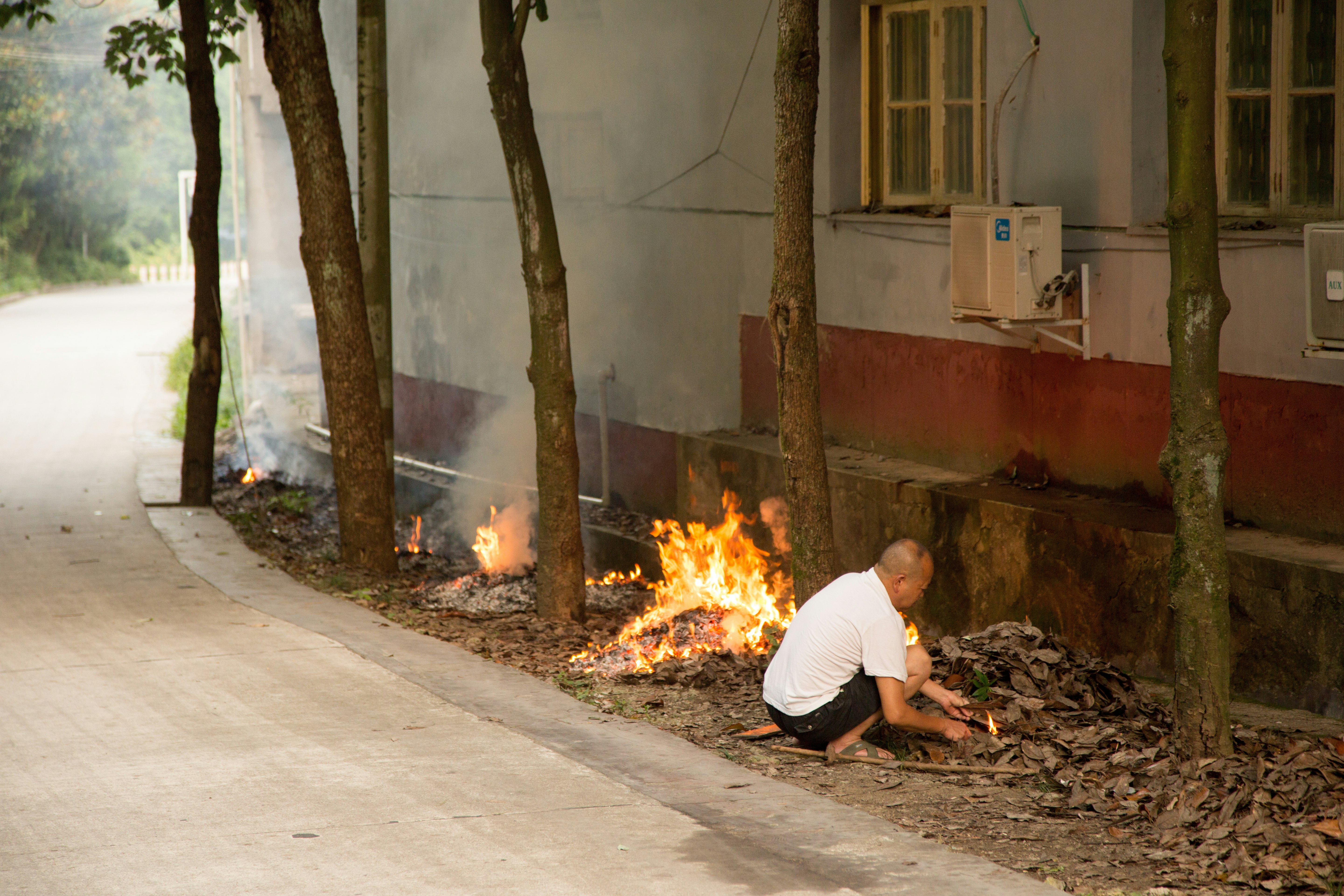man in white t-shirt and black pants sitting on ground near burning woods during daytime